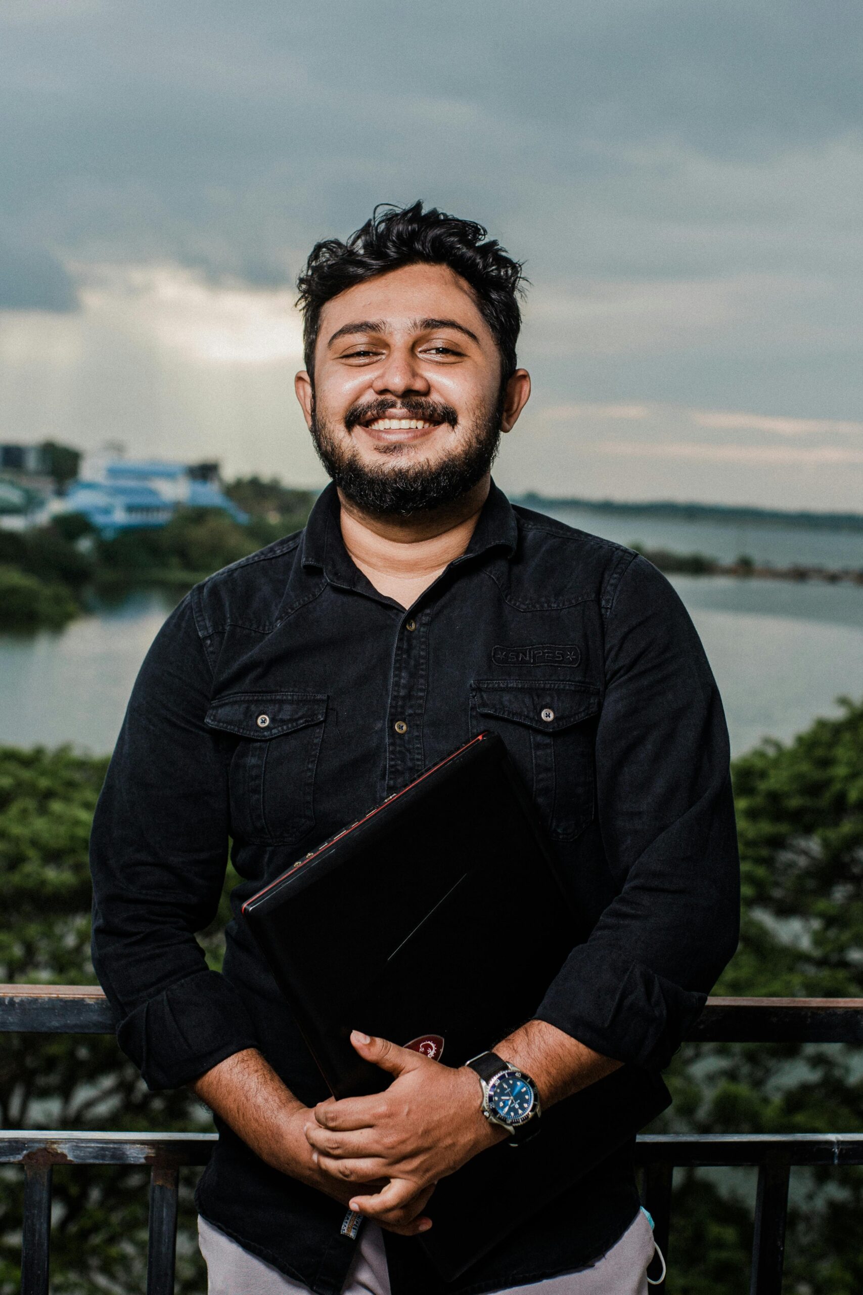 A cheerful man with a beard holds a laptop outdoors with a scenic view in the background.